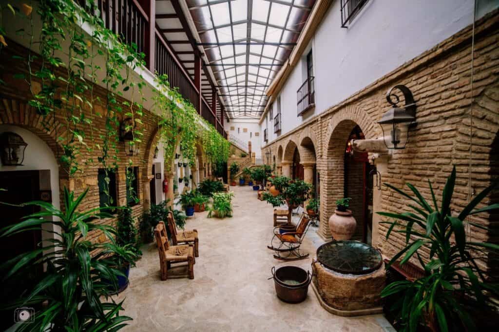 A very cozy, earthy patio, that is long and skinny, has bricked archways along the sides, and white walls above it, ivy hanging down, potted plants, and mutliple chairs and places to sit.