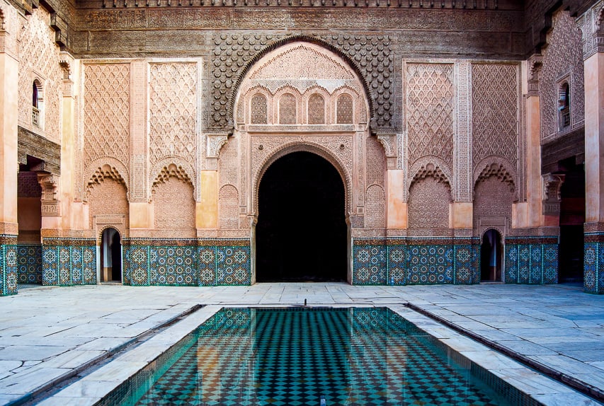 A view of the Madrasa ben Youssef, with a basin of water in the middle, stone flooring, large arches on the walls, and tiles and carved stucco adorning everything.
