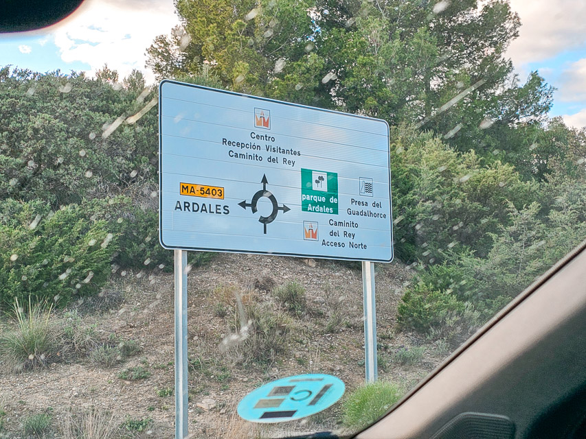 A white sign displays a black circle with arrows to indicate a roundabout and information is displayed about the various destinations down each path.