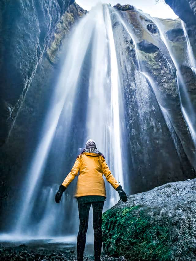 A woman stands in front of a big waterfall that comes down over a very tall cliff wall.