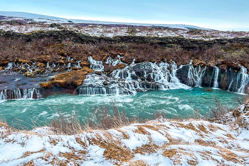 A lot of waterfalls cascading down rocks into a blue river with snow fields all around.