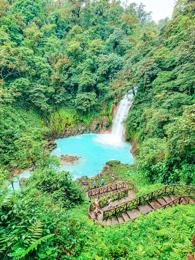 View of the waterfall going down into a bright blue pool from high up, with staircase leading down.