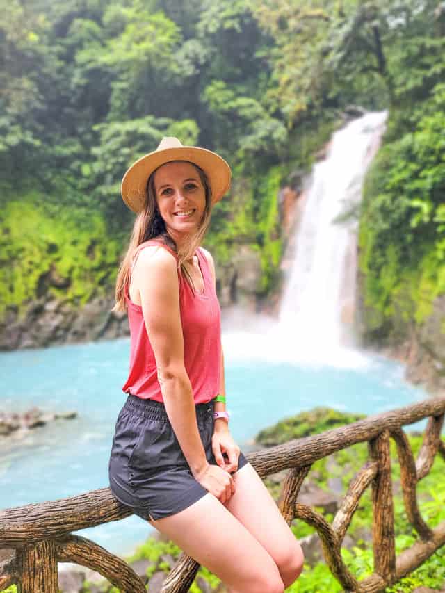 Woman standing in front of waterfall, with trees in the background.