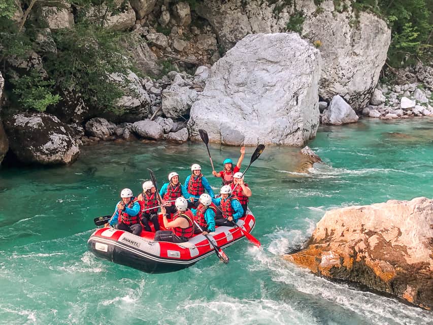 A group of 9 people in a bright red raft smile on the Soča River which is a beautiful blue, clear, color.