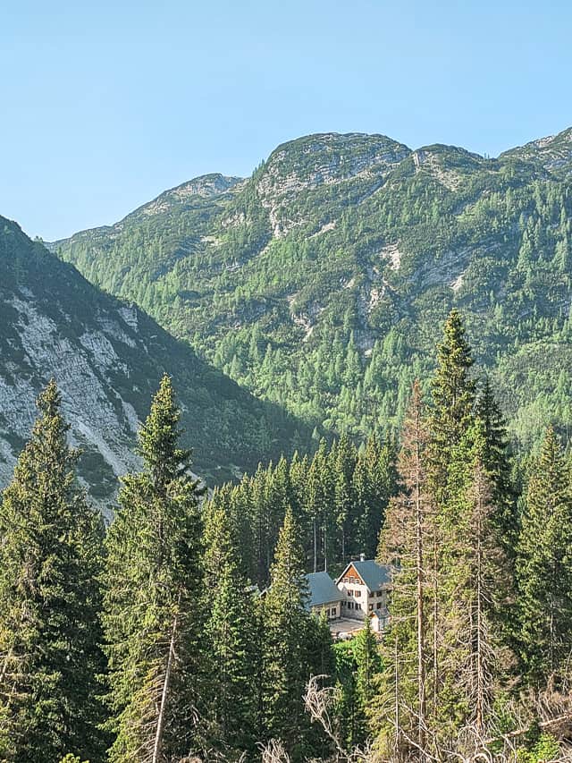 A small wood hut tucked between the pine trees at the base of a large mountain hit with sun rays lighting up the top.