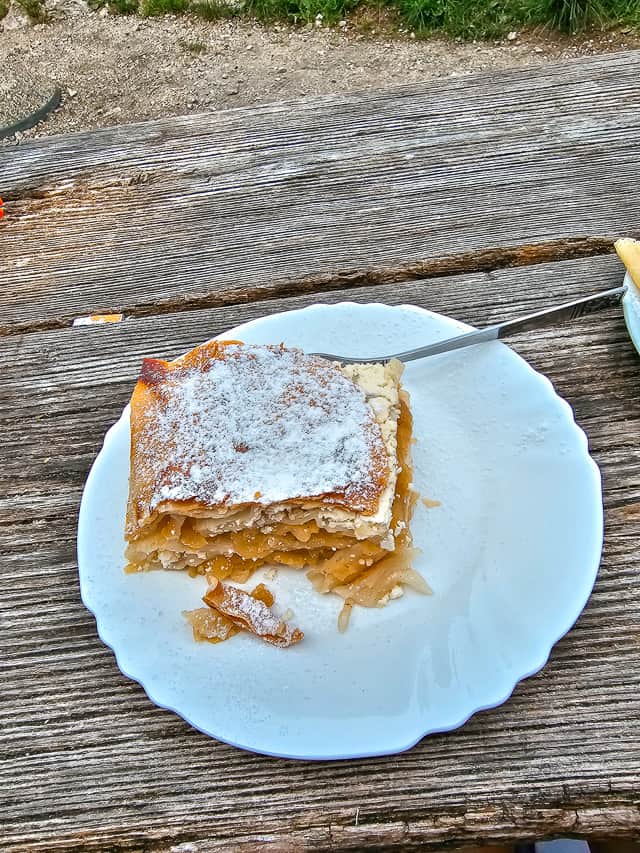 A homemade apple strudel with multiple layers and powdered sugar on a white plate on a brown wood table.