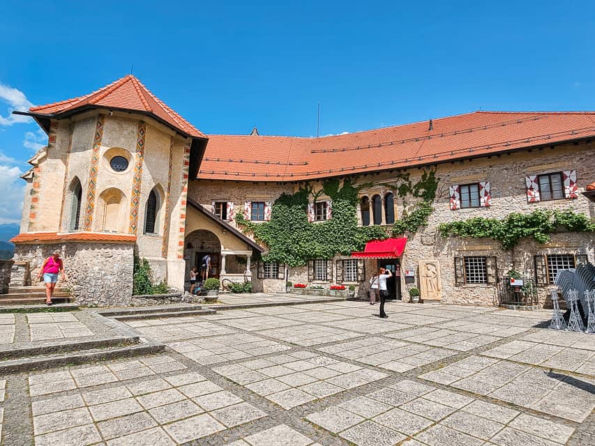 People walk around a courtyard with a castle like stone building with gray walls and a red roof. This is one of many incredible things to do on the Lake Bled island.