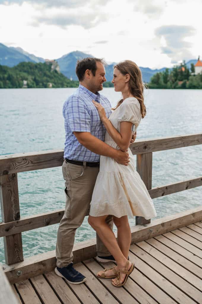 A man and woman hold each other on a boardwalk looking over Lake Bled and mountains in the background.