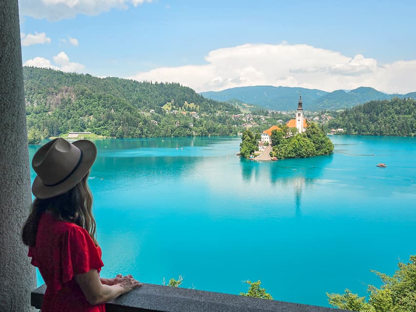 A woman looks at an island on Lake Bled surrounded by blue water in the lake with mountains in the distance.