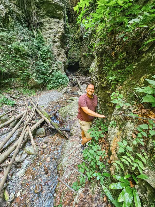A man smiles holding on to a wire and standing on poles driven into the rocks. with a small stream and wood logs below him.