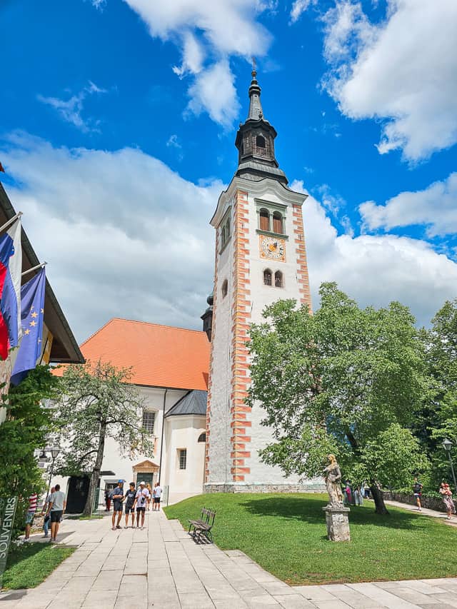 A tall steeple stands in the middle of a courtyard with a tree, and then flags to the other side.