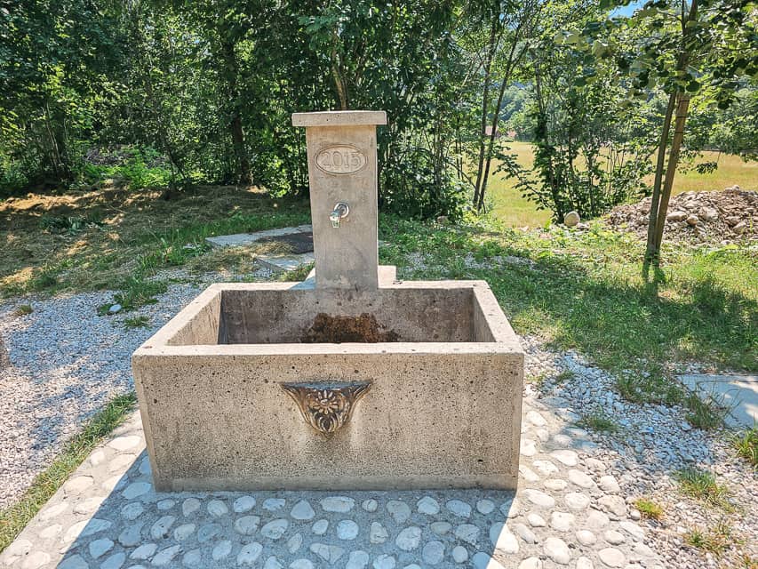 A stone water spigot with an engraving saying 2015 on a gravel road and a field with trees next to it.