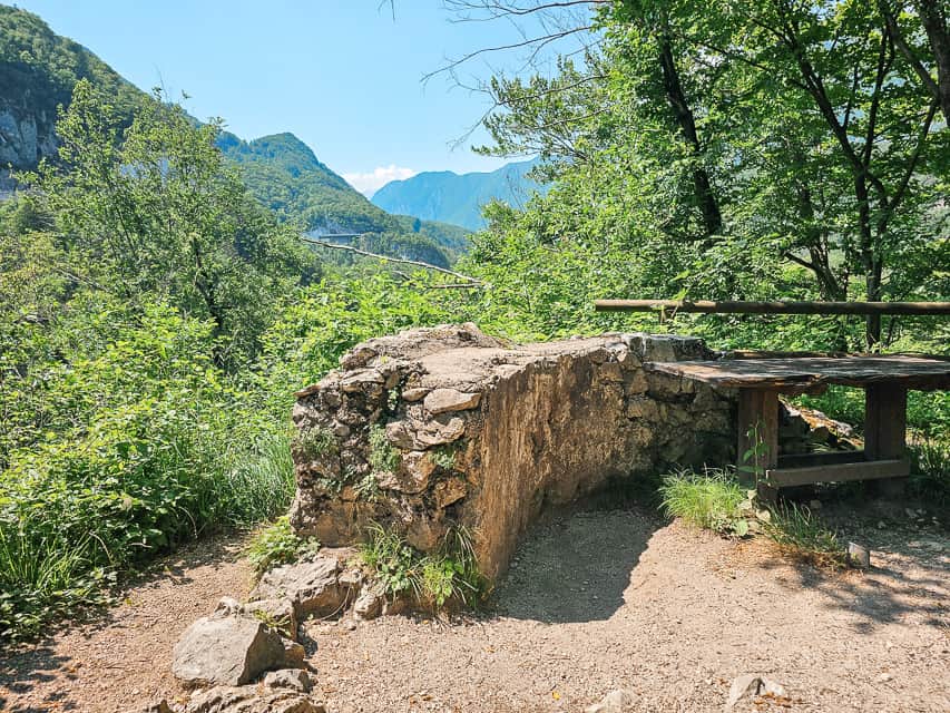 A small stone wall follows the curve of the dirt trail with large green mountains in the distance. Part of the WWI fortifications in the Soca River Valley.