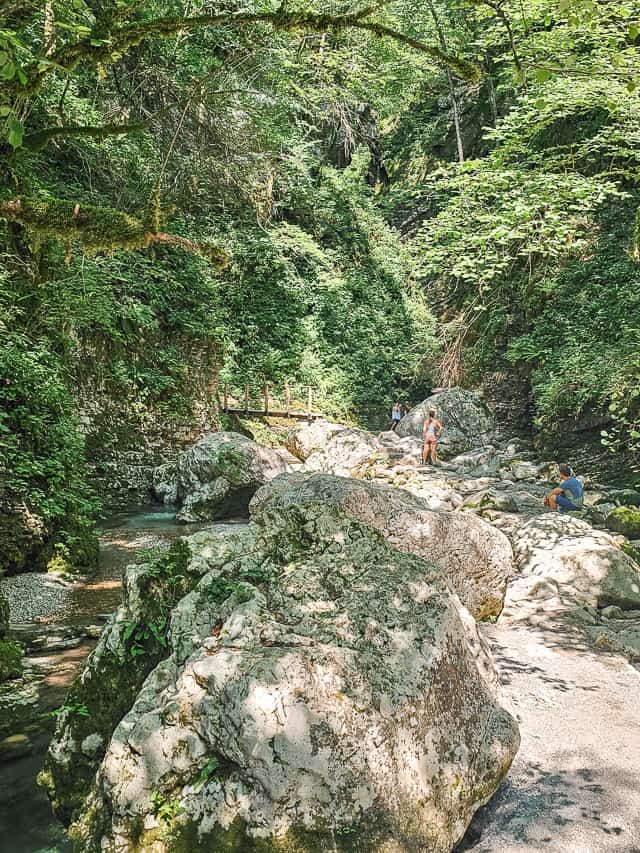 A few people stand on the rocks next to the stream with trees and a wood bridge in the distance along the trail.