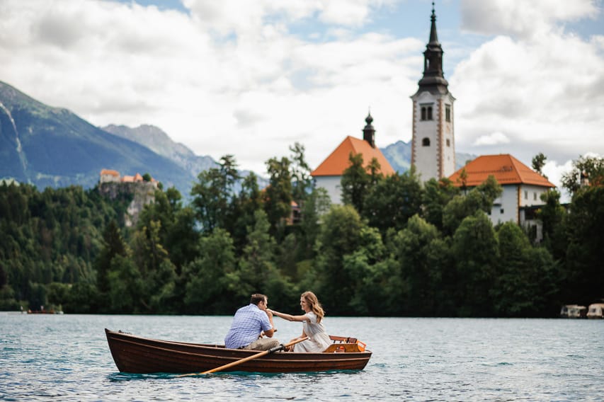 A man and a woman sit in a rowboat on Lake Bled, with the church steeple on Bled Island in the background, and some mountains behind them.