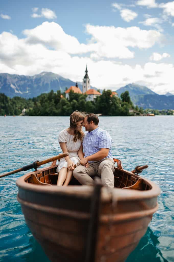A romantic couple snuggle noses on a rowboat with an island and mountains in the background.