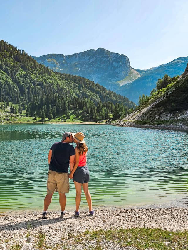 A man and woman kiss on the edge of a blue and green lake with pine trees and large mountains on the other side of the lake.
