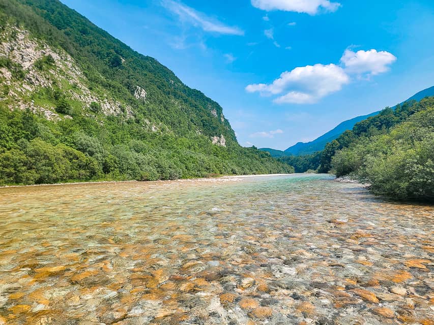 A view from the shore of a beach on the Soča River with crystal clear water and mountains in the background.