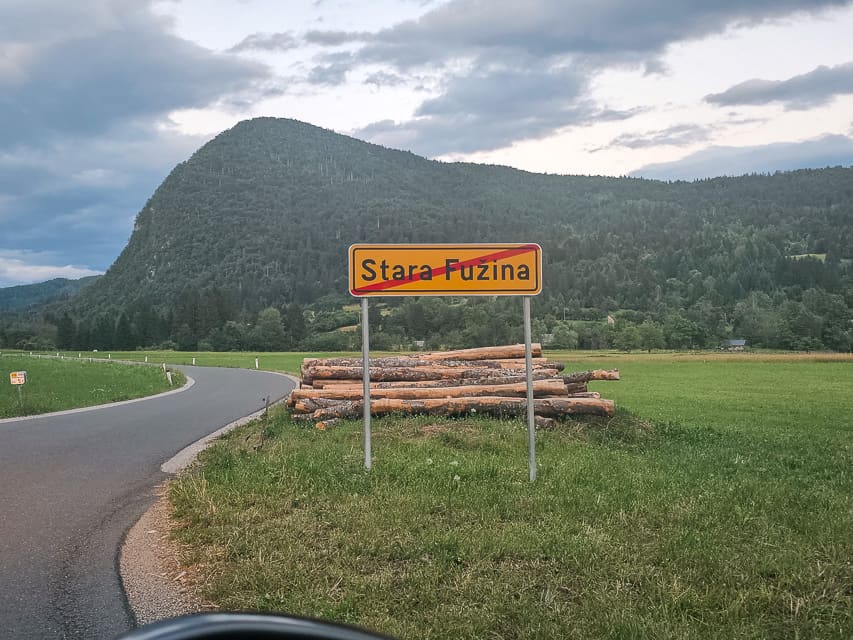 A yellow rectangular sign with a vilage name, and a red slash through it, with hills in the background.