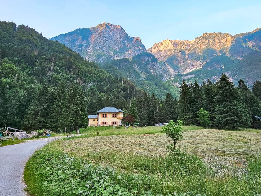 A cement and gravel road leading through a grass field winding up to the sheer mountains with the sun just starting to hit the top of the mountain peaks.