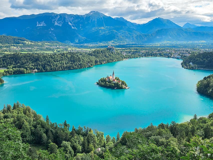 A lake with a island in the middle with a church steeple on it is, is surrounded by trees and mountains.