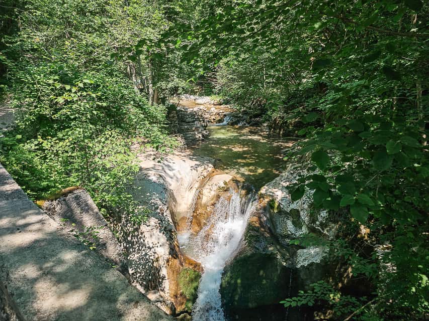 A stone bridge with a river cascading down some rocks with ferns and trees all around.