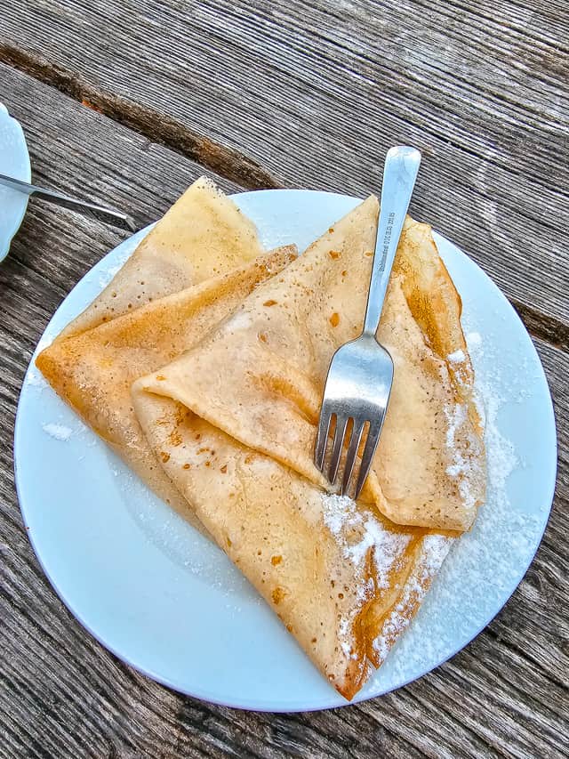 2 yellow crepes coated with powder sugar with on a white plate on a dark bown wood table.