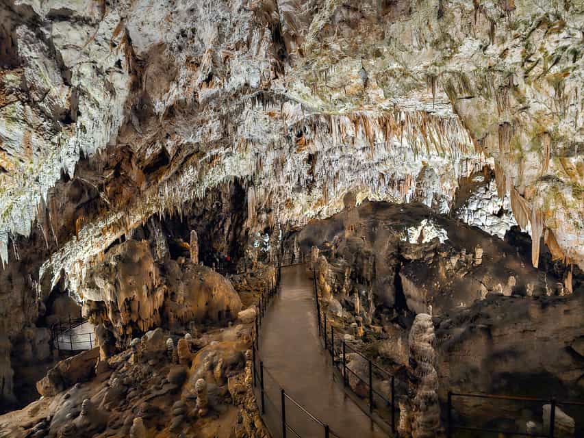 A large cavern with a large rock overhang goes over a rock and metal path throughout the stalagmites and stalactites.