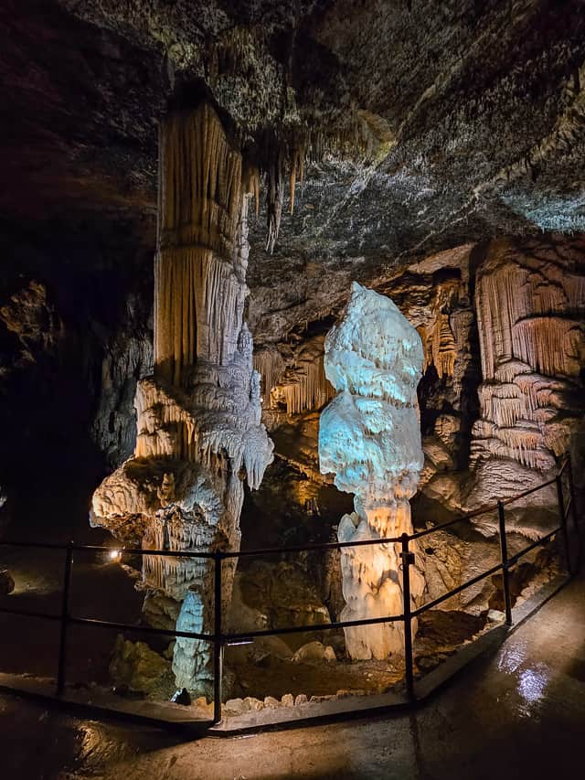 A lit path through a cavern shows rock pillars with horizontal and vertical pillars carved into the pillars.