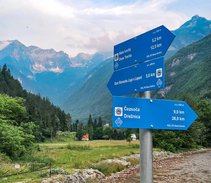 Signs point to different places with mountains in the distance and trees all around it.