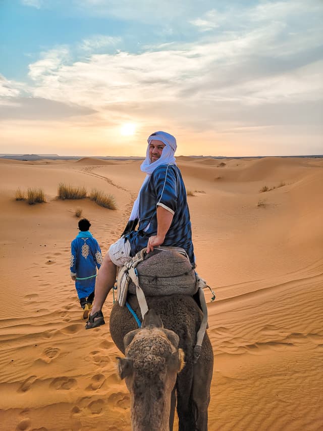 A man looks back on the back of a camel with a tour guide walking on the sand dunes in the Sahara desert. This is one of the best and highlight things to do in the Sahara desert.