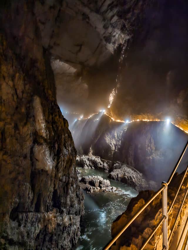 A lighted path follows a cliff through a huge cavern with a rushing river in the bottom of the gorge.