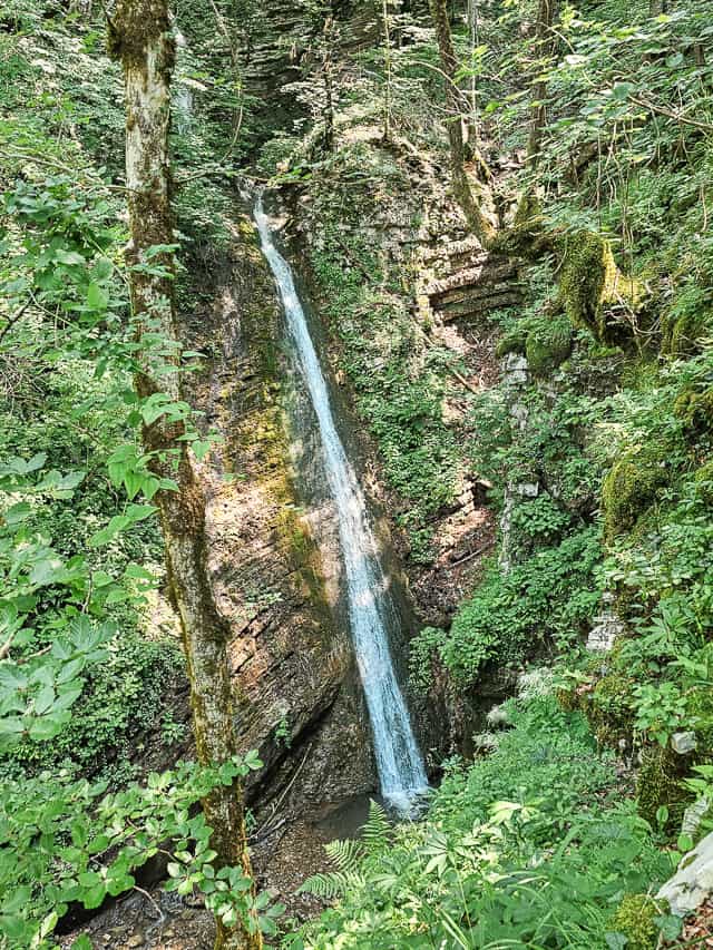 A large waterfall slides of mossy rocks into a small pool with gravel as a base.