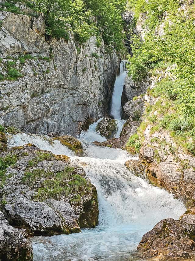 A waterfall sandwiched between rocks goes into a rushing powerful river. This is a unique slovenian waterfall.