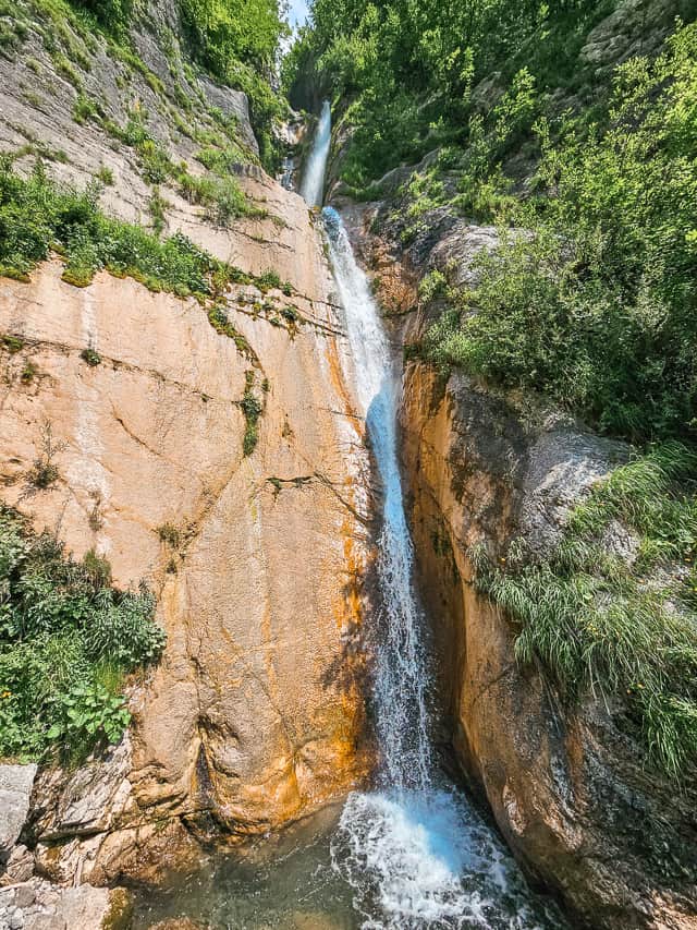A tall waterfall falls into a pool with lots of greenery growing on cliffs around it.