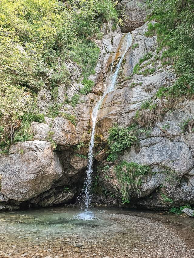 A waterfall goes down into a crystal clear pool, with many trees surrounding it. This is a very pretty slovenian waterfall.