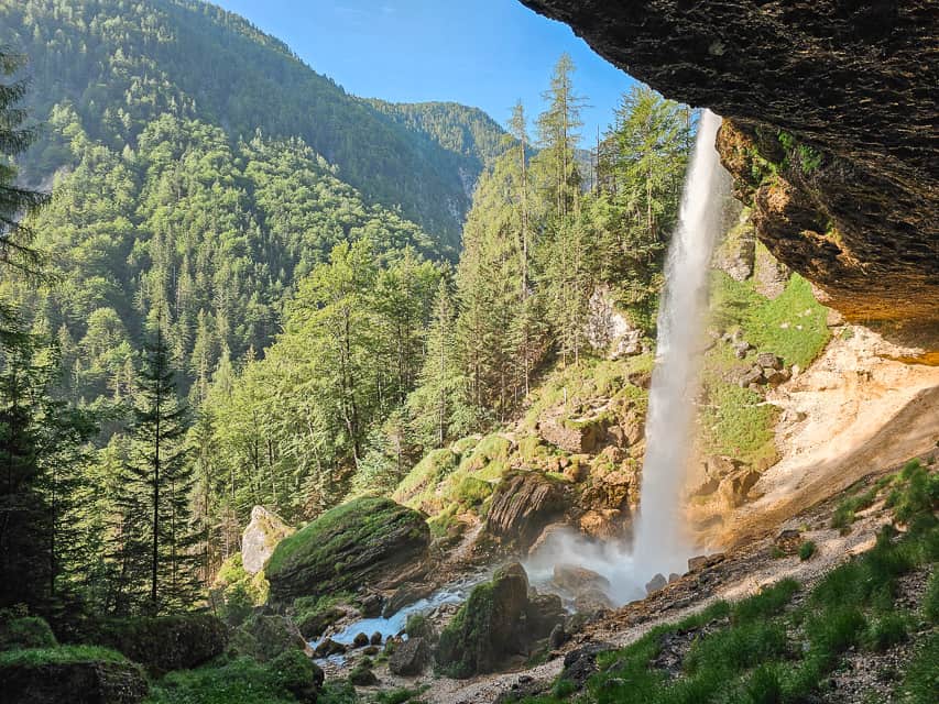 A waterfall goes down over a cliff and free falls for a second and then drops down into a river. You can see the path behind the waterfall that visitors can walk on.