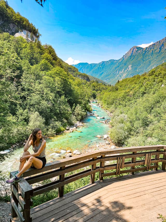 A woman sits on a platform, looking at the turquoise river that runs underneath, with mountains in the distance, and green trees all around it.
