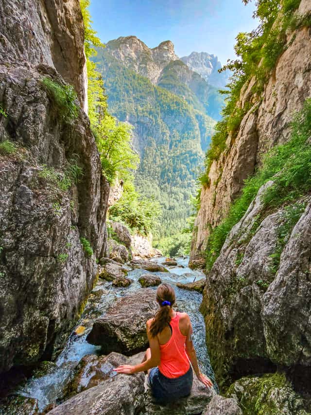 A woman sits on a rock while a river rushes past her legs, with tall cliff walls on both sides, and tall mountains in the distance.