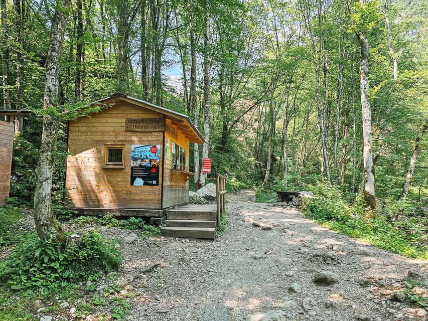 A brown wood booth stands on the side of the trail to the Kozjak Waterfall with the dirt and gravel trail continuing down through the grove of trees.