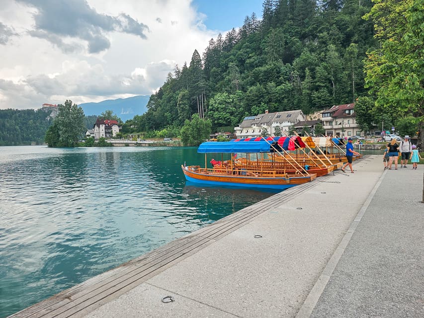 A sidewalk parrallel to Lake Bled with pletnas lined up against the sidewalk and trees along the lakeside.