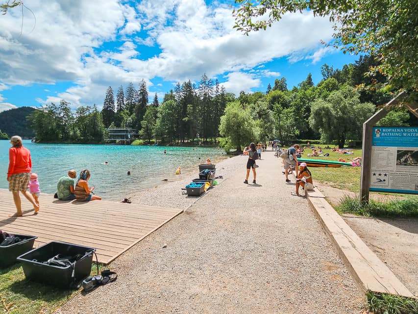 A gravel sidewalk right to to a beach with a small boardwalk with lots of trees around the edge of the lake.