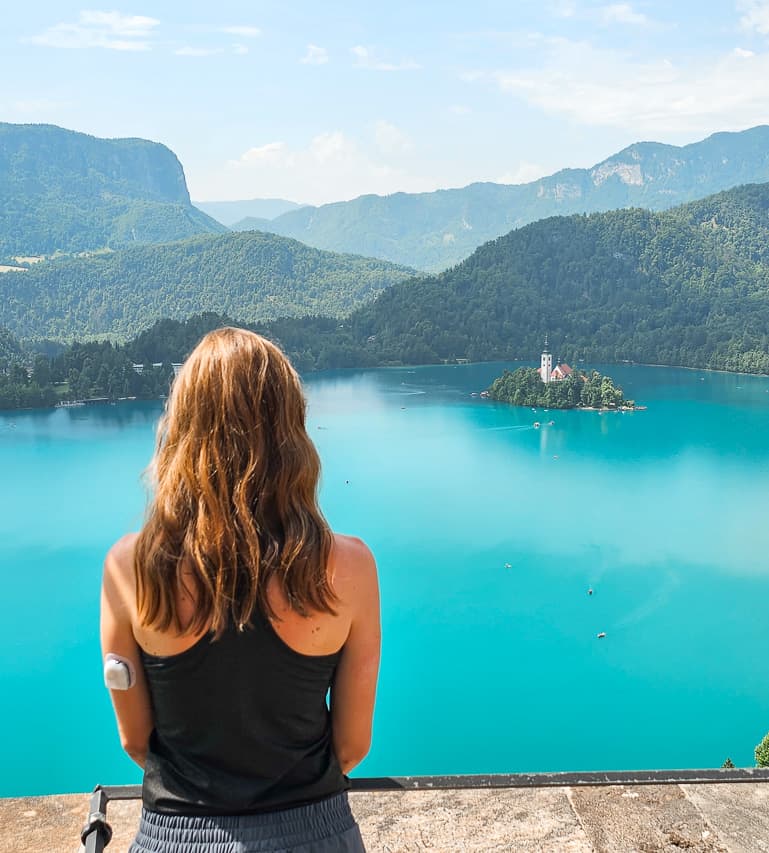 A woman stands at a viewpoint looking at a crystal blue lake and small island with hills overlooking the background .