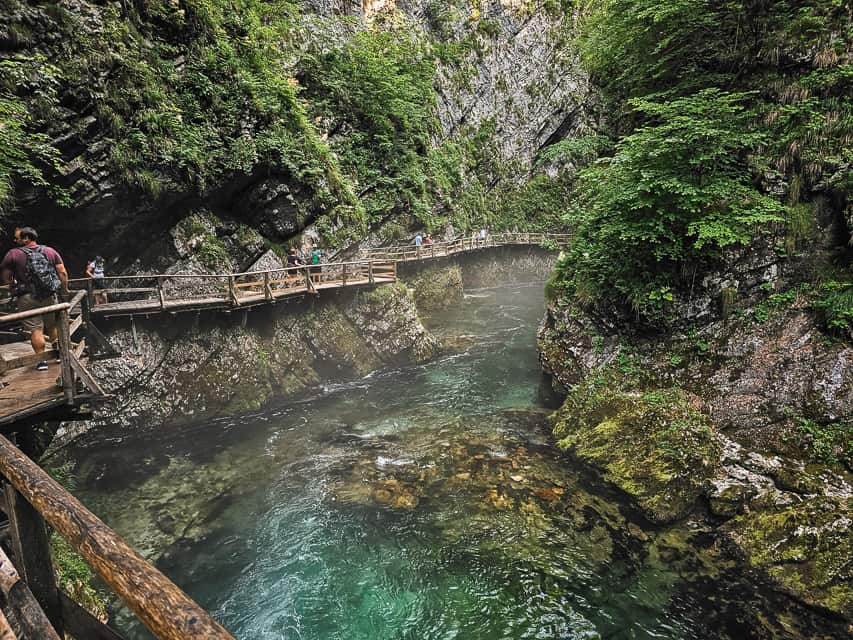 People walk on a wood boardwalk on the side of rock walls with a river cascading beside them.