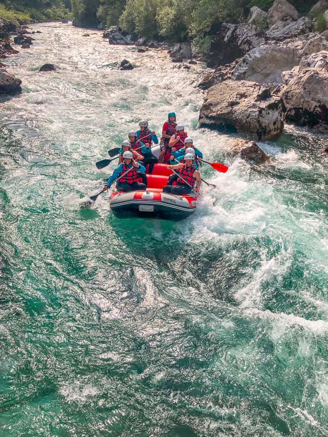 A group of 9 people on a red raft paddle down a white water rapid on the Soča River.