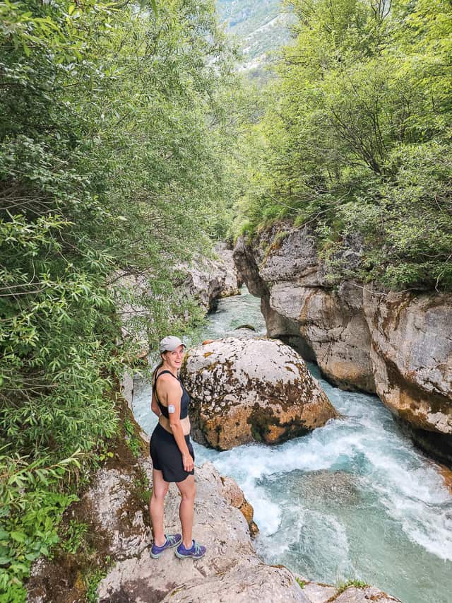 A woman stands on grey rocks overlooking the blue Soca River through a rock gorge.