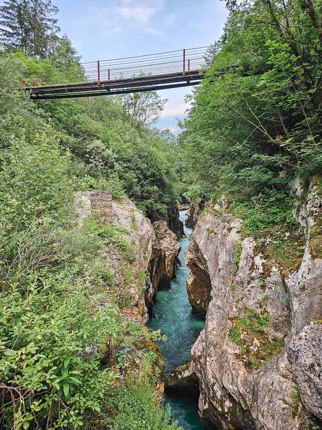 The Soca River runs through 2 slot canyon walls with a metal and wood bridge over it.