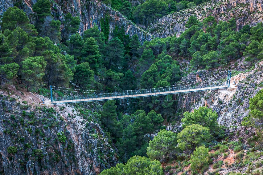 A metal and wood bridge connects 2 sides of a grey and rock cliff.
