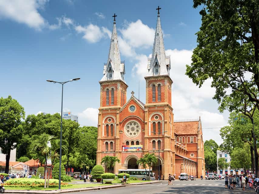 A brown stone church with 2 steeples in the middle of a busy street with trees and busses.