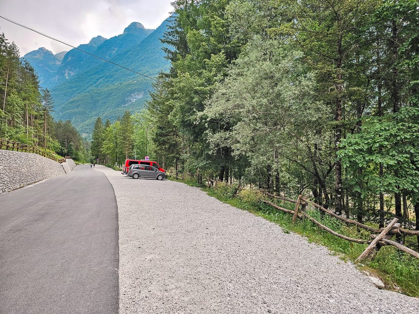 A gravel parking lot on the right side at the trailhead for the Soca River and a concrete street on the left side with trees everywhere and a mountain range in the background.